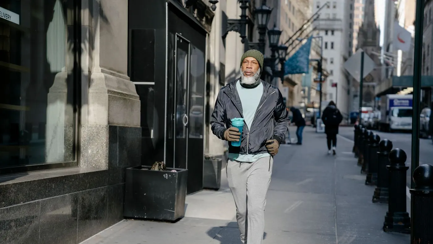 Hombre mayor con gorro y ropa deportiva que camina por una acera soleada con un vaso térmico en la mano: imagen para la guía sobre la andropausia