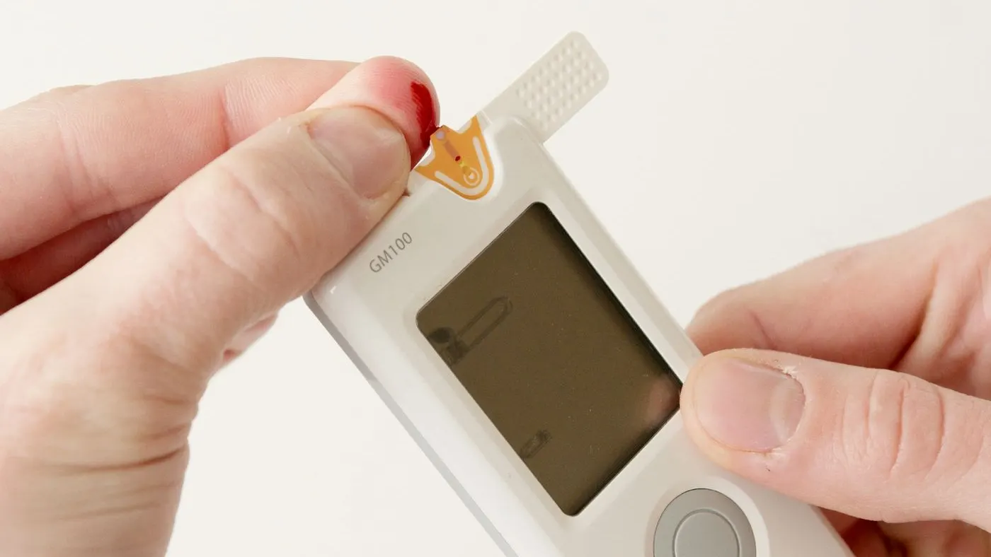 Manos que sostienen un glucómetro blanco con una tira reactiva y una gota de sangre en la yema del dedo: imagen para la guía sobre diabetes y disfunción eréctil