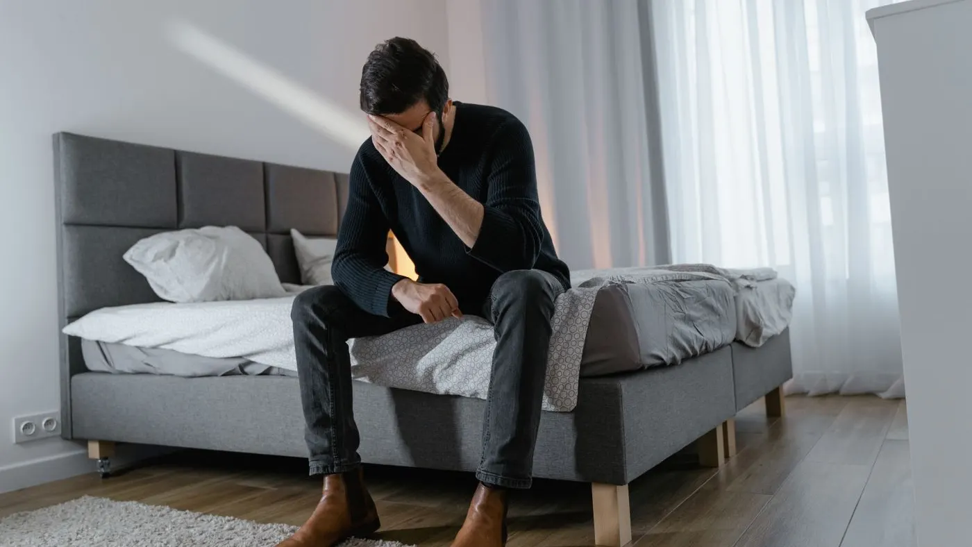 Hombre de mediana edad con suéter negro sentado en el borde de una cama gris, con una mano en la frente y la mirada baja, en un cuarto con cortinas claras: imagen para la guía sobre la disfunción eréctil psicológica
