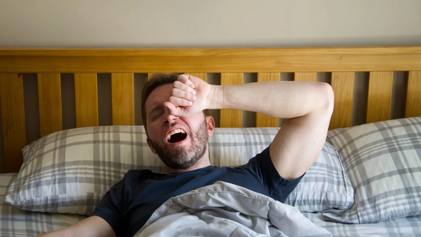 Hombre de barba que bosteza y se frota los ojos al despertar en una cama con ropa a cuadros: imagen para la guía sobre la erección matutina