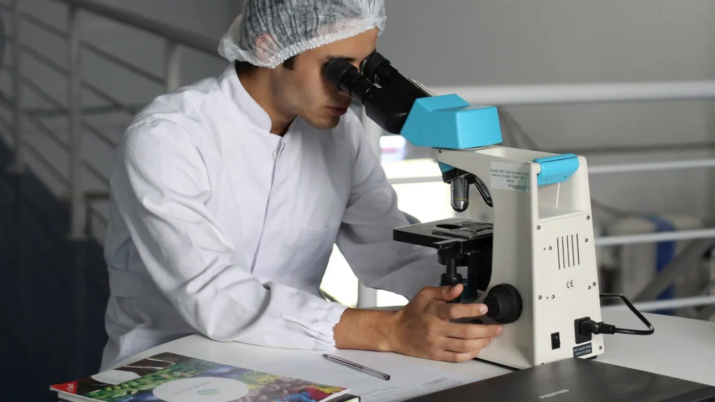 Técnico de laboratorio con bata blanca y gorro mirando por un microscopio sobre una mesa de trabajo: imagen para el espermograma
