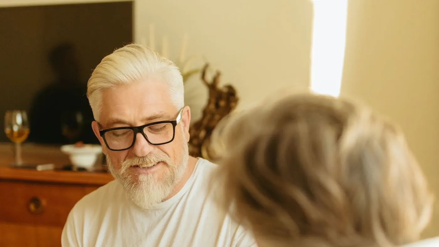 Hombre de pelo y barba canosos, con gafas de marco negro y camiseta blanca, sentado en la sala de una casa mientras conversa con otra persona: imagen para la guía sobre hipertensión y disfunción eréctil