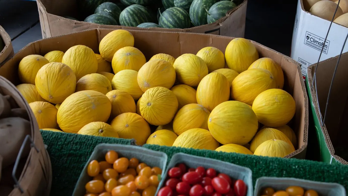 Cajas de tomates cherry rojos y amarillos frente a una pila de melones amarillos y sandías en un puesto de mercado: imagen para la guía del licopeno