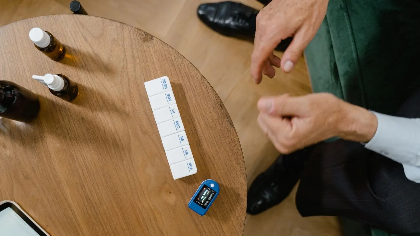 Mesa redonda de madera con un pastillero semanal, frascos pequeños de medicina y un oxímetro, y las manos de un hombre con camisa blanca junto a la mesa: imagen para la guía sobre medicamentos que pueden causar disfunción eréctil