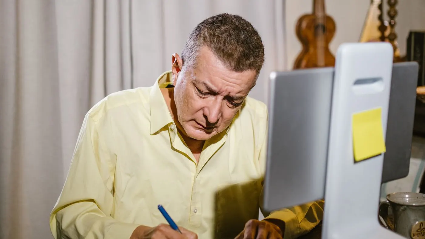 Hombre de camisa amarilla clara que anota algo en un cuaderno sobre un escritorio, con una pantalla de computador al lado: imagen para anotar desde cuándo se sienten los síntomas de la próstata inflamada