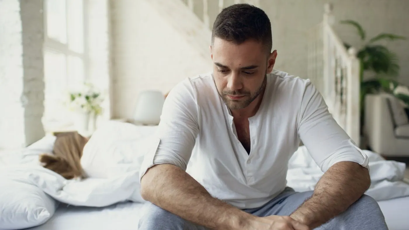 Hombre de camiseta clara sentado al borde de la cama con la mirada baja en un cuarto luminoso, con una persona acostada detrás: imagen para la guía sobre la testosterona baja