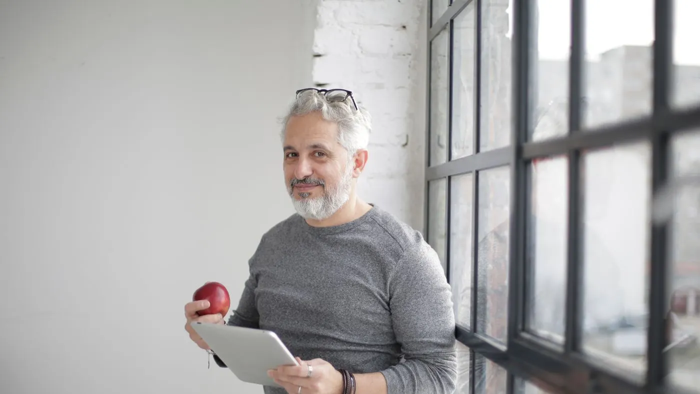 Hombre de mediana edad con barba gris, tableta y una manzana roja en la mano, junto a un ventanal: imagen para la guía sobre el varicocele