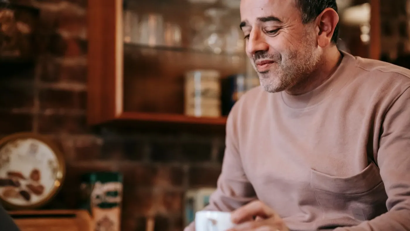 Hombre de mediana edad con barba corta sonríe frente a una vitrina de cocina mientras sostiene una taza de café: imagen para la guía sobre disfunción eréctil e impotencia
